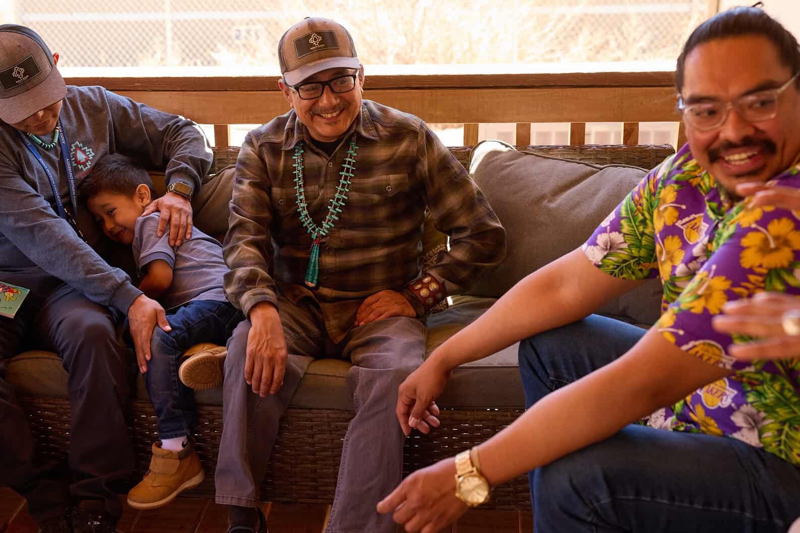 Four men and a child sit together on an outdoor couch; one man comforts the child, while the others talk and smile.