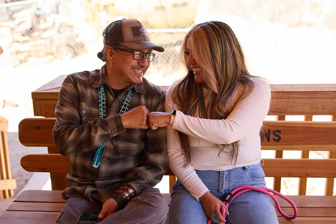 Randy and Joi Lynch sit on a wooden bench, smiling and giving each other a fist bump.