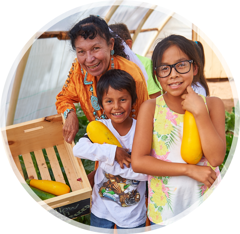 A joyful older woman and two children smile while holding large yellow squash in a greenhouse. The scene is vibrant and full of warmth.