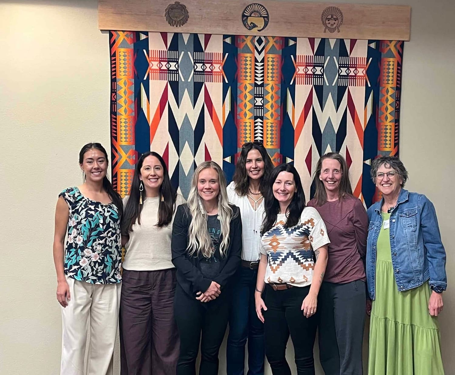 Seven women stand in a line and smile in front of a colorful, geometric-patterned textile hanging on a wall.
