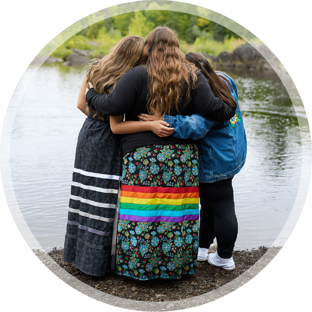 Three young women in ribbon skirts hugging each other on the shore of a river.
