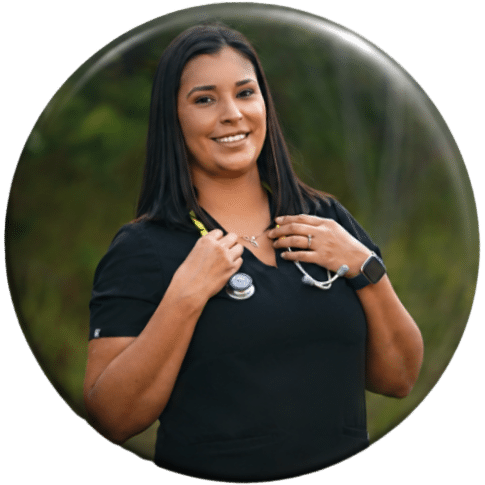 A healthcare professional with a stethoscope around her neck smiles, standing outdoors in a black scrub top, with greenery in the background.