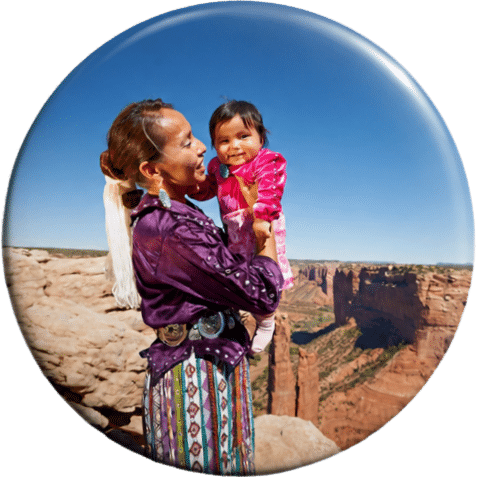 Navajo woman in traditional dress holds up a smiling baby against a backdrop of Spider Rock in Canyon de Chelly.